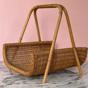 May include: A vintage wicker magazine rack with a curved base and a handle. The rack is made of woven light brown wicker and has four wooden legs. The handle is also made of wood and is arched. The background is a solid pink color.