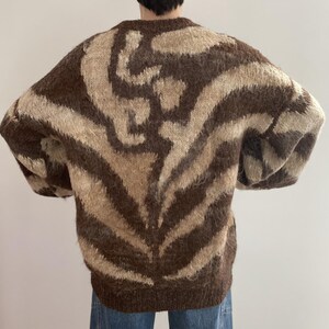 Pu&ograve; includere: Un maglione marrone e beige fantasia. Il maglione ha un girocollo e maniche lunghe. Il motivo &egrave; un disegno a vortice marrone e beige. Il maglione &egrave; realizzato in un materiale morbido e soffice.