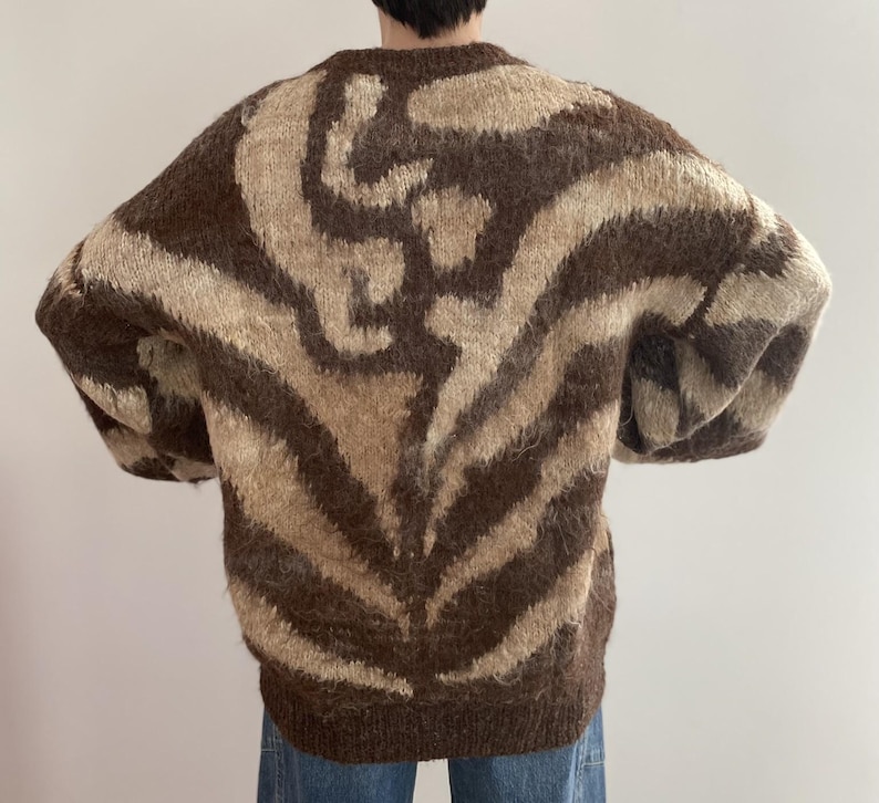 Pu&ograve; includere: Un maglione marrone e beige fantasia. Il maglione ha un girocollo e maniche lunghe. Il motivo &egrave; un disegno a vortice marrone e beige. Il maglione &egrave; realizzato in un materiale morbido e soffice.