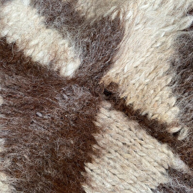 Pu&ograve; includere: Primo piano di un tessuto intrecciato e strutturato con un motivo marrone, beige e bianco sporco. Il materiale sembra spesso e soffice, con fili visibili e una superficie leggermente irregolare. Il design presenta un motivo geometrico.