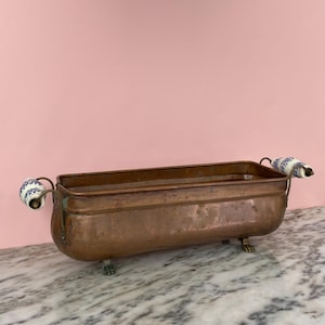 May include: A rectangular copper planter with blue and white ceramic handles and decorative feet. The planter has a weathered appearance. The handles are curved. The planter is approximately 30 cm long, suitable for a variety of plants.