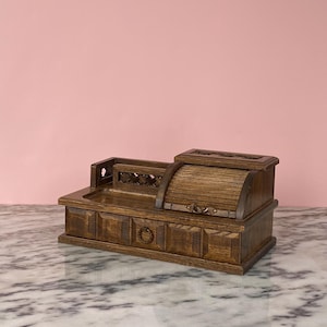 Puede incluir: Un organizador de escritorio de madera vintage con una sección enrollable y un cajón pequeño. El organizador es de madera oscura y presenta tallas decorativas y un tirador de latón. El fondo es de color rosa liso.