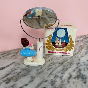 May include: A vintage Doll-Up Mirror with a chrome frame and a decorative base featuring a figurine in a blue dress. The original box is visible, with the text "DOLL-UP MIRROR" and a graphic of a woman.