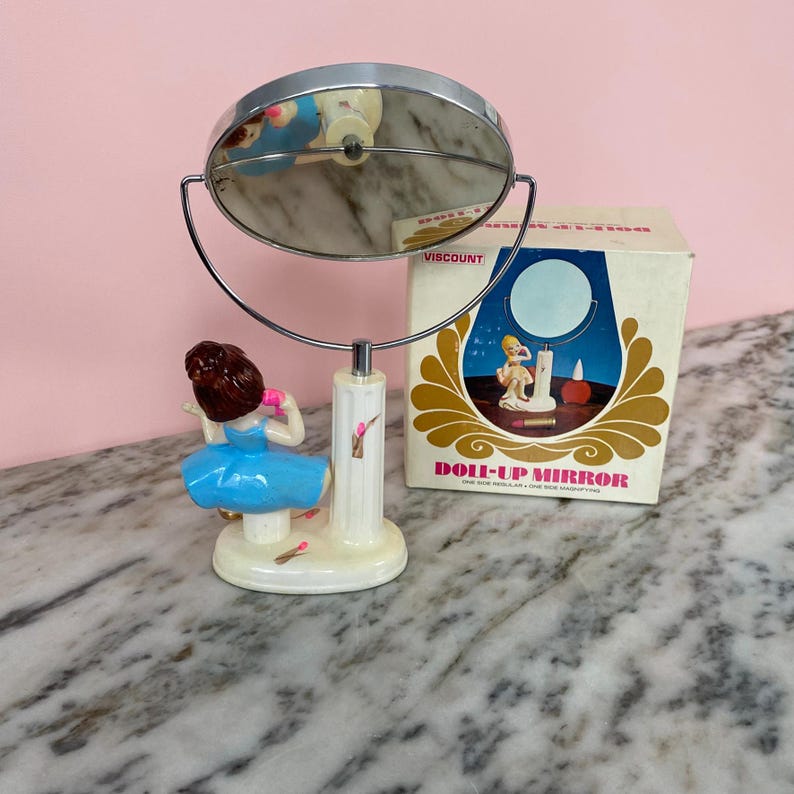 May include: A vintage Doll-Up Mirror with a chrome frame and a decorative base featuring a figurine in a blue dress. The original box is visible, with the text "DOLL-UP MIRROR" and a graphic of a woman.