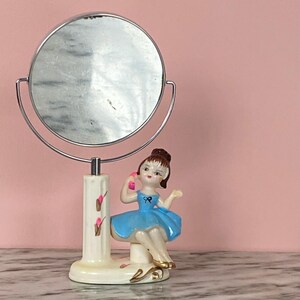 May include: A vintage vanity mirror with a chrome frame and a decorative porcelain figurine. The figurine is of a girl in a blue dress, holding a pink telephone. The base is off-white with pink floral accents.