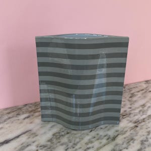 May include: A grey ceramic vase featuring horizontal wavy stripes in a darker shade of grey. The vase has a rectangular shape with a slightly curved base and top, set against a pink background.