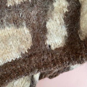 Pu&ograve; includere: Primo piano di un tessuto strutturato lavorato a mano a maglia nei toni del marrone e del crema. Il motivo presenta forme irregolari e una trama spessa e sfocata, che suggerisce un capo o un accessorio caldo e accogliente.