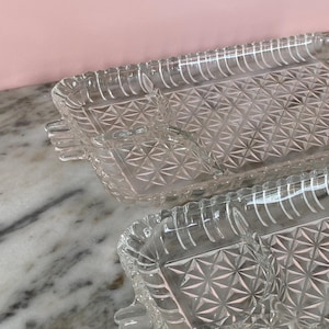 May include: Two clear glass serving trays with a diamond pattern and scalloped edges. Each tray has a rectangular shape with a small, attached compartment. The trays are on a marble surface.