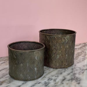 Peut inclure: Deux jardinières antiques en métal avec un motif tissé. Les jardinières cylindriques ont une finition sombre et vieillie. La plus petite mesure environ 15 cm de diamètre, et la plus grande environ 20 cm. Elles sont posées sur une surface en marbre.