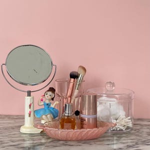May include: A vanity scene with a round, double-sided mirror, a small ballerina figurine, makeup brushes, perfume, skincare products, and a glass jar with cotton swabs. The items are arranged on a pink, leaf-shaped dish.