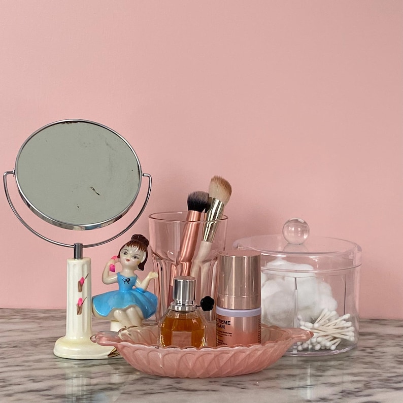 May include: A vanity scene with a round, double-sided mirror, a small ballerina figurine, makeup brushes, perfume, skincare products, and a glass jar with cotton swabs. The items are arranged on a pink, leaf-shaped dish.