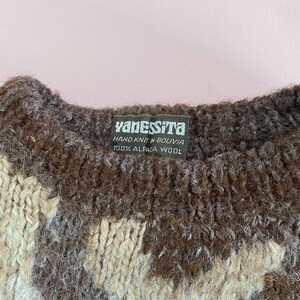 Pu&ograve; includere: Primo piano di un maglione lavorato a mano con un'etichetta che recita "YanesSita, Lavorato a mano in Bolivia, 100% Lana di alpaca". Il maglione presenta un motivo di colori marrone, beige e crema.