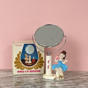 May include: A vintage vanity mirror with a chrome frame and a decorative base featuring a porcelain figurine of a ballerina in a blue dress. A box with the text "Doli-Up Mirror" is also present.