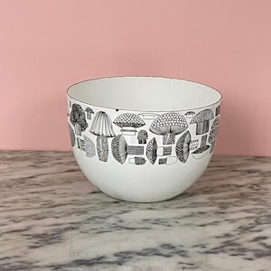 Puede incluir: Cuenco de cerámica blanco con un patrón de setas en blanco y negro alrededor del borde. El cuenco tiene forma redondeada y se apoya sobre una superficie de mármol sobre un fondo rosa. El diseño de setas presenta varios estilos.