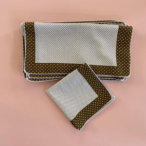 May include: A set of white and brown polka dot napkins. The napkins have a white center with small brown polka dots and a brown border. The napkins are stacked on a pink surface.