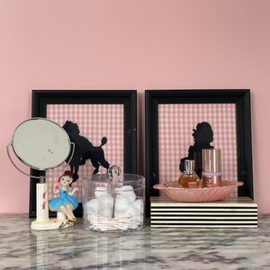 May include: A vanity scene with a black framed mirror, a porcelain ballerina figurine, and a clear jar filled with cotton balls and swabs. Two framed poodle silhouettes hang on a pink gingham background. A pink leaf-shaped dish holds perfume and skincare products.
