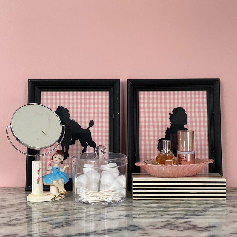 May include: A vanity scene with a black framed mirror, a porcelain ballerina figurine, and a clear jar filled with cotton balls and swabs. Two framed poodle silhouettes hang on a pink gingham background. A pink leaf-shaped dish holds perfume and skincare products.
