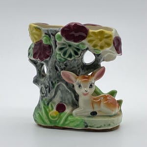 May include: A ceramic planter shaped like a tree stump with a resting fawn at the base. The tree trunk is grey with textured bark, adorned with yellow and maroon flowers. The fawn is light brown and white with painted black eyes and eyelashes.