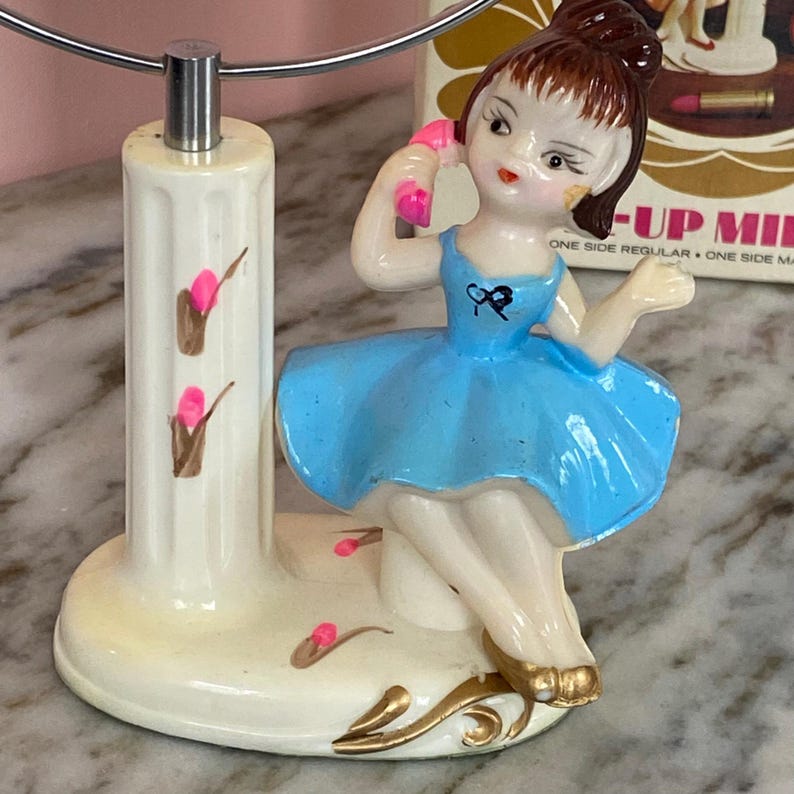May include: A vintage vanity mirror with a decorative base featuring a porcelain figurine of a girl in a blue dress. The figurine is holding a pink telephone. The base is cream-colored with pink and brown floral accents.