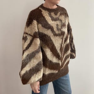 Pu&ograve; includere: Un maglione fantasia marrone e beige con scollo tondo e maniche lunghe. Il maglione ha un design a zebratura ed &egrave; realizzato in un materiale morbido e strutturato. Le maniche sono ampie e il maglione &egrave; oversize.