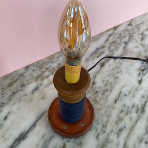 May include: A small, handmade table lamp with a warm, amber-colored bulb. The lamp features a wooden base with a layered design of brown, blue, and orange. A yellow paper ring sits between the bulb and the wooden base. A black cord extends from the lamp.