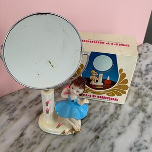 May include: Vintage Doll-Up Mirror with a round, silver-toned mirror and a ceramic base featuring a ballerina figurine in a blue dress. The mirror is accompanied by its original box, which has the words "Doll-Up Mirror" printed on it.