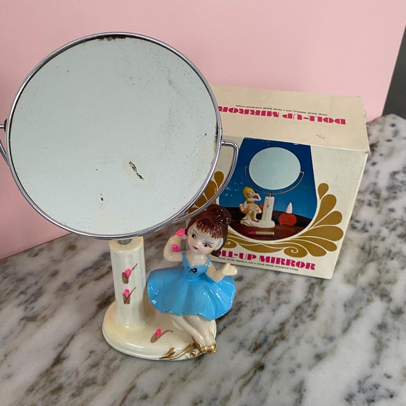 May include: Vintage Doll-Up Mirror with a round, silver-toned mirror and a ceramic base featuring a ballerina figurine in a blue dress. The mirror is accompanied by its original box, which has the words "Doll-Up Mirror" printed on it.