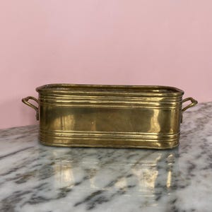 Puede incluir: Una jardinera rectangular de latón con dos asas pequeñas en los laterales. La jardinera tiene crestas horizontales y una superficie brillante y reflectante. El fondo es una pared rosa claro.