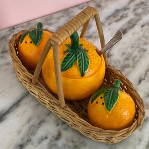 May include: A ceramic orange-shaped condiment set in a woven basket. The set includes a lidded bowl with a spoon, and two salt or pepper shakers. The oranges are bright orange with green leaves. The basket has a handle.