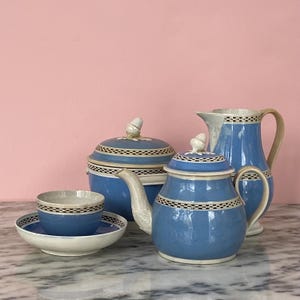 Puede incluir: Un juego de té de cerámica azul y blanco con tetera, jarra de crema, azucarero, taza y platillo. El juego tiene un patrón de cuadros blancos y negros y remates en forma de bellota. El fondo es rosa.