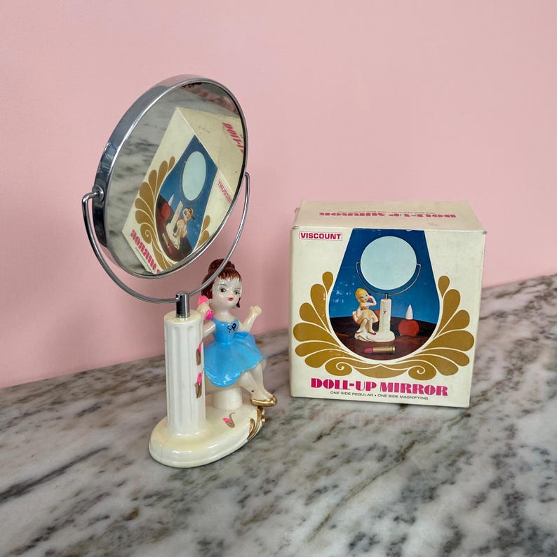 May include: A vintage vanity mirror with a chrome frame and a decorative base featuring a porcelain doll in a blue dress. The mirror is next to its original box, which reads "DOLL-UP MIRROR". The box has a gold and blue design.