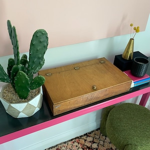 May include: A rectangular wooden box with a hinged lid and a small round knob, positioned next to a green cactus in a geometric patterned pot. The box features visible metal hinges along the back edge and a natural wood grain finish throughout.