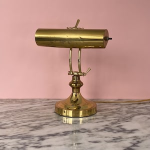 May include: A classic brass desk lamp featuring a cylindrical shade and an adjustable arm. The lamp has a circular base and a switch. It sits on a marble surface, with a pink backdrop.