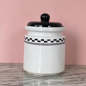 Puede incluir: Un tarro de cerámica blanco con tapa y pomo negros. El tarro presenta un patrón de cuadros blancos y negros en el centro. Un recipiente decorativo para guardar en la cocina.