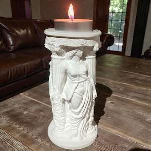 May include: A white, classical-style candle holder with a burning tea light candle. The holder features a sculpted female figure and architectural details. The candle flame is orange and yellow. The holder is on a wooden table.