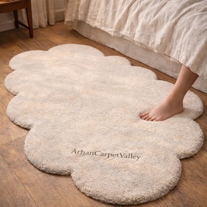 Op de afbeelding: Een crèmekleurig, wolk-vormig vloerkleed met een zacht, getextureerd oppervlak. Het vloerkleed ligt op een houten vloer bij een bed. De tekst "ArhanCarpetValley" is op het vloerkleed gedrukt.