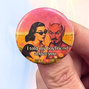 May include: A red and white button with a retro illustration of a woman whispering to a devil with horns. The text on the button reads "I told my boyfriend about you."