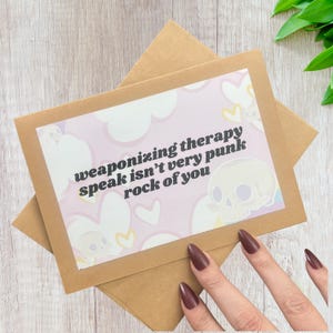 Puede incluir: Una tarjeta de felicitación con el texto "weaponizing therapy speak isn't very punk rock of you" sobre un fondo rosa con ilustraciones de calaveras y nubes. La tarjeta está dentro de un sobre marrón.