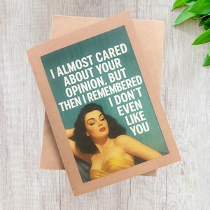 Puede incluir: Una tarjeta de felicitación con una imagen retro de una mujer con un vestido amarillo. La tarjeta tiene el texto "I ALMOST CARED ABOUT YOUR OPINION, BUT THEN I REMEMBERED I DON'T EVEN LIKE YOU". La tarjeta está sobre un fondo marrón.