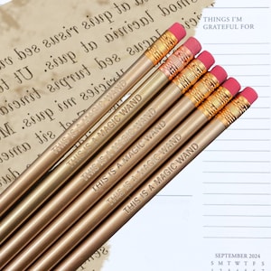 May include: Five gold pencils with pink erasers. Each pencil has the text "This is a magic wand" engraved on the side.