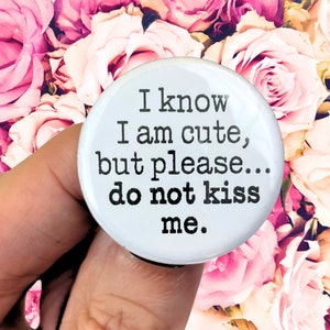 May include: White button with black text that reads "I know I am cute, but please... do not kiss me."