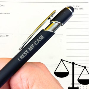 以下が含まれることがあります： シルバーのクリップとスタイラスチップが付いた黒いペン。ペンには「I rest my case」という文字が刻まれています。背景には黒い正義の天秤があります。