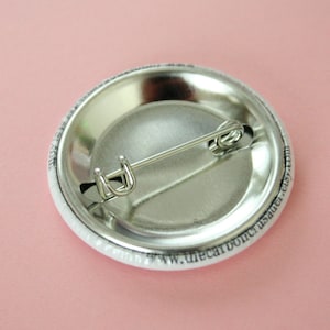 May include: The back of a white button with a silver pin and black text that reads "www.thecarboncrust.etsy.com".