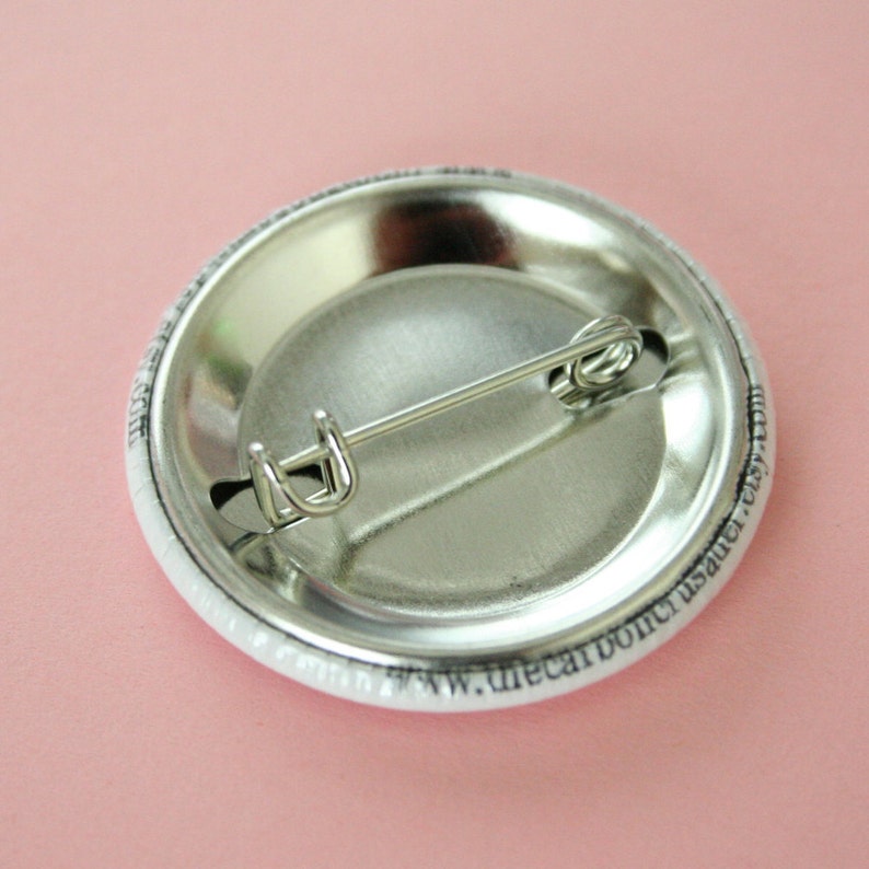 May include: The back of a white button with a silver pin and black text that reads "www.thecarboncrust.etsy.com".