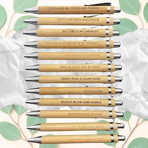 May include: A collection of bamboo pens with silver accents. Each pen is engraved with an inspirational quote, including "With God All Things Are Possible" and "Faith Can Move Mountains". The pens are arranged on a light background.
