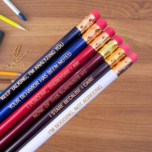 May include: Set of six pencils with gold-colored accents and funny sayings printed on them. The sayings are: "Keep talking, I'm analyzing you", "Your behavior has been noted", "I perceive, therefore I am", "None of this surprises me", "I stare because I care", and "I'm nodding, not agreeing".