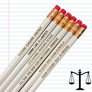 Op de afbeelding: Zes witte potloden met gouden letters en rode gummen. De potloden hebben zinnen als "Future Esquire Energy" en "Lucky Bar Exam Pencil". Een zwart symbool van de weegschaal van de rechtvaardigheid staat rechtsonder.