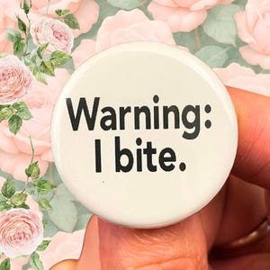 May include: A round, off-white button with the text "Warning: I bite." in black. The button is held up against a background of pink roses and green leaves.