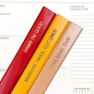 May include: Three pencils with humorous phrases printed on them. The red pencil says "DAMN I'M GOOD", the yellow pencil says "MEASURE TWICE, CUT ONCE", and the natural wood pencil says "I CAN MAKE THAT". The pencils are set against a white calendar page.