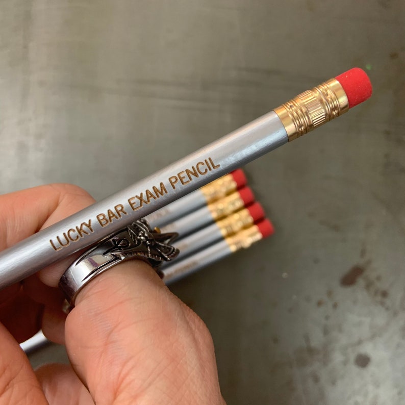 May include: A silver pencil with a red eraser and gold accents. The pencil is engraved with the text "Lucky Bar Exam Pencil".